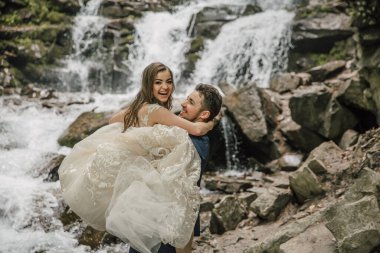 Şelalenin yanında mutlu bir gelin ve damadın portresi. Damat gelini kollarında tutuyor. Doğada düğün fotoğrafı çekimi. Gelin ve damadın ormanında fotoğraf çekimi..