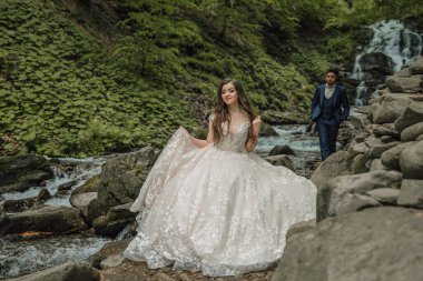 Dağ nehrinin yakınında bir düğün çifti. Ön planda odaklanmış durumda. Damat ve gelin. Doğada düğün fotoğrafı çekimi. Gelin ve damadın ormanında fotoğraf çekimi..