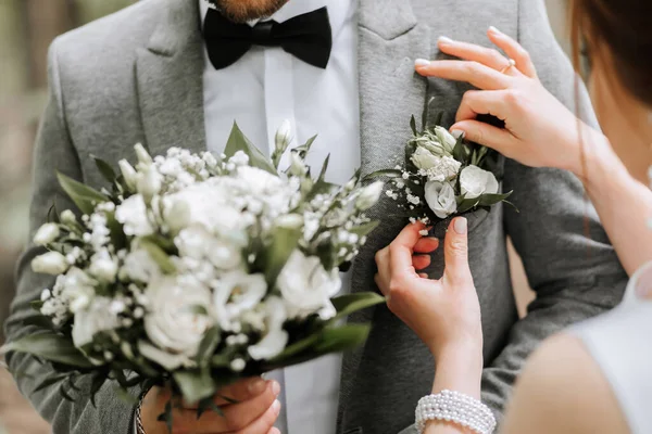 The groom is holding a bouquet of flowers. The bride attaches the boutonniere to the groom\'s jacket. Photo details