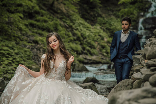 Wedding couple near a mountain river. She is in the foreground in focus. Groom and bride. Wedding photo session in nature. Photo session in the forest of the bride and groom.