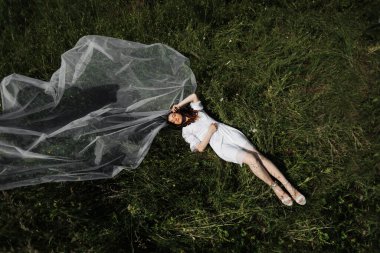 Gelinin doğadaki sabahı. Gelin sabahlık ve uzun bir peçeyle yeşil çimlerin üzerinde uzanıyor ve güneşin sıcaklığının tadını çıkarıyor. Geniş açı fotoğrafı.