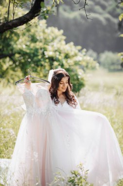 Uzun kıvırcık saçlı güzel bir gelin narin elbisesini tutar ve bahçede ona hayran kalır. Gelinin sabahı. Doğal makyaj.