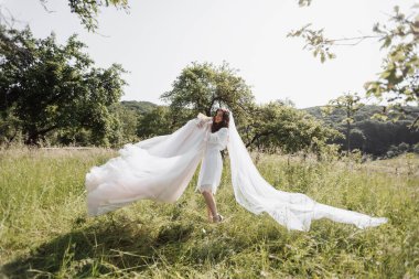 Beyaz bornozlu mutlu gelin gelinlikle bahçede dans ediyor. Yeşil çimlerin üzerine kolayca düşen bir peçe. Gelinin bahçede geçirdiği sabah. Kırsal tarzda. Bahar düğünü. Parlak gün ışığı