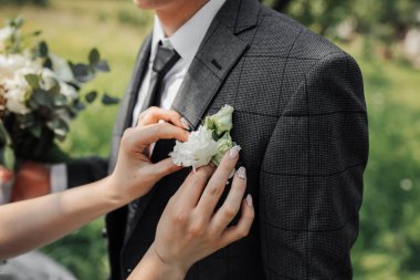Gelin bir yaka çiçeği tutuyor. Gelin ellerinin düğün töreninde damadın ceketine yaka çiçeği dikerken çekilmiş fotoğrafı. Damat bir buket tutuyor. Fransız manikürü. Düğün detayları