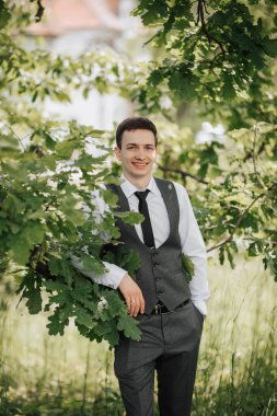 Mutlu şık damat kameraya bakarak, bir ağaç dalına yaslanarak samimi bir şekilde gülümsüyor. Damadın portresi.