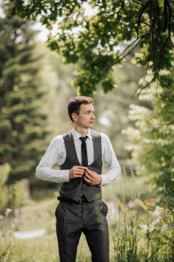 Mutlu şık damat ceketinin düğmelerini ilikliyor ve başka tarafa bakıyor. Yelek giymiş damat. Damadın portresi. Bahar düğünü