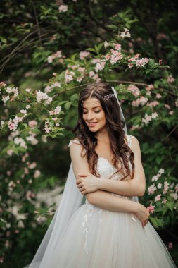 Şık bir elbise içinde uzun kıvırcık saçlı güzel bir gelin, gülümsüyor, düğün buketini tutuyor, lense bakıyor. Gelinin portresi. Bahar düğünü. Doğal makyaj.