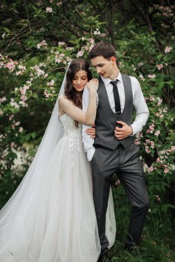 Mutlu genç çift. Düğün portresi. Gelin nazikçe damada sarıldı. Genç gelin ve damat kameraya bakarken şefkatle kucaklaşıyorlar. Düğün buketi. Bahar düğünü