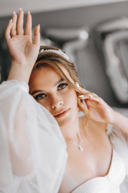 Beyaz elbiseli güzel kıvırcık saçlı bir gelin, bir odada kolları olan güzel bir elbise içinde dururken bir fotoğrafçıya poz veriyor. Düğün fotoğrafçılığı, yakın plan portre.