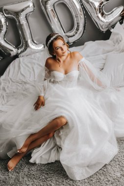 Beyaz elbiseli güzel kıvırcık saçlı bir gelin, kolları olan güzel bir elbise içinde bir yatakta otururken bir fotoğrafçıya poz verir. Düğün fotoğrafçılığı, yakın plan portre, şık saç stili..