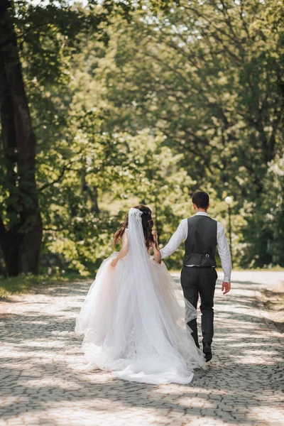新郎和新娘在花园里 春天的婚礼在公园举行 快乐的新婚夫妇在公园里奔跑 又时髦又漂亮照片来自后面 公主礼服 — 图库照片