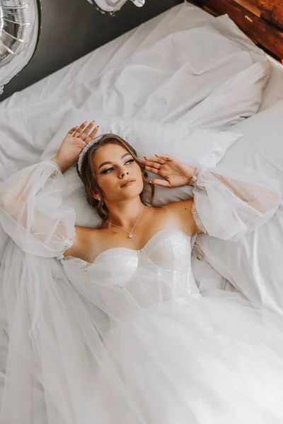 Красивая Кудрявая Каштановая Невеста Белом Платье Позирует Фотографу Лежащему Кровати — стоковое фото