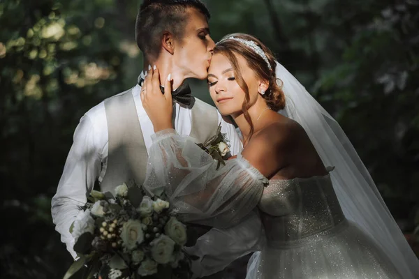 結婚式の日に愛の美しいカップルの肖像画 太陽の光の下で公園を散歩 素晴らしいキスと花束で花嫁と新郎の抱擁 — ストック写真