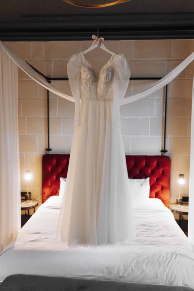一件完美的婚纱 衣架上有一条郁郁葱葱的裙子 床边有一块红色的床头板和一张白色的床单 还有一块装饰新娘房间的烤饼 — 图库照片