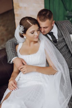 Gelin ve damat yeşil pencerelerin arkasında bir kanepede oturuyorlar. Şık gelin ve damat. Düğün fotoğrafı. kolay atmosfer