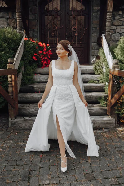 Romantic full-length portrait of a brown-haired woman in a white dress with a bouquet standing near the wooden gate of the church entrance