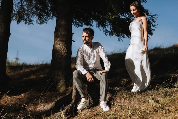 stock image Stylish model couple in the mountains in summer. A young girl in a white silk dress walks along the slope against the background of the forest and mountain peaks.