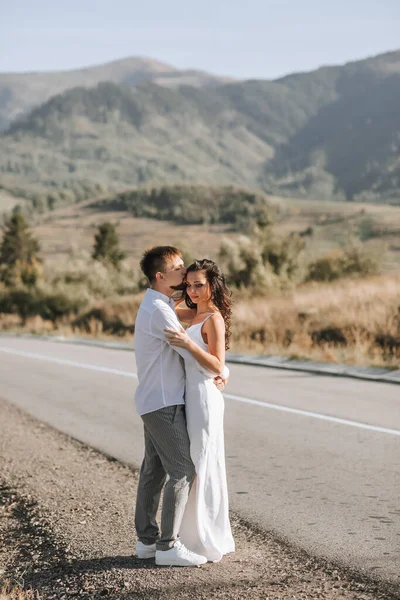 白いシャツを着たスタイリッシュな新郎と白いドレスを着たかわいいブルネットの花嫁は 森や山の背景を背景に道路の側面に抱いています 新婚の結婚式の肖像画 — ストック写真
