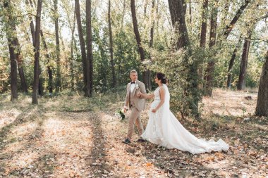 Düğün fotoğrafı. Gelin ve damat ormanda el ele tutuşup birbirlerine bakıyorlar. Damat bir buket tutuyor. Birbirine aşık bir çift. Yaz ışığı. Gölgeler