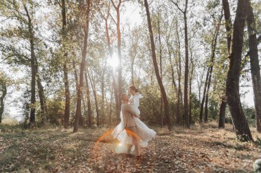 Ormanda mutlu gelin ve damadın portresi. Damat gelini kollarında tutuyor, onunla dans ediyor. Havada uzun bir peçe var. Fotoğraftaki bir güneş ışığı. Geniş açı fotoğrafı