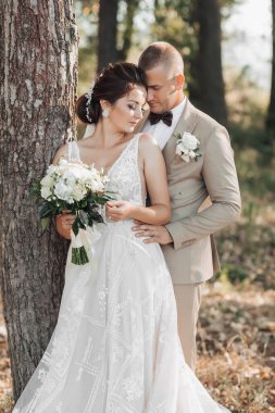 Doğada düğün fotoğrafı. Gelin ve damat bir ağacın yanında duruyorlar, damat arkadan sevgilisine sarılıyor, gelin ise gözleri kapalı bir buket tutuyor. Portre. Yaz düğünü