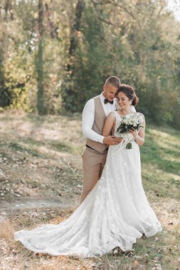 Doğada düğün fotoğrafı. Damat sevgilisine arkadan sarılıyor ve ona bakıyor, gelin bir buket tutuyor ve yana bakıyor. Elbisenin uzun treni. Portre. Yaz düğünü.