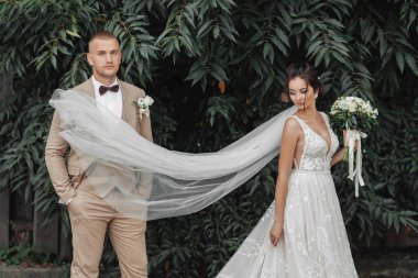 Yeşil arka planda düğün fotoğrafı. Gelin ve damat yeşil çalıların yanında duruyorlar. Tam boy şık bir damat. Havaya atılan uzun peçeli güzel bir gelin. Düğün portresi