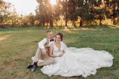 Gelin ve damadın düğün portresi. Gelin ve damat yeşil çimlerin üzerinde oturup poz veriyorlar. Gelin ve damadın arkasında, güzel güneş büyük Noel ağaçlarının arasından parlıyor..