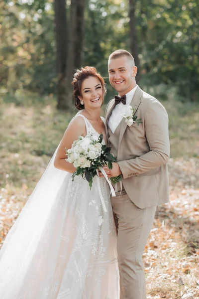 Wedding portrait. The bride and groom are standing in the forest hugging and looking at the camera, both holding a wedding bouquet. Long dress with train. Light suit of the groom. Beautiful makeup