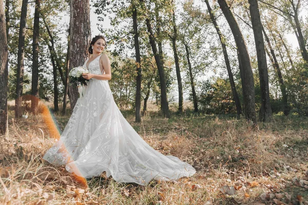 自然の中で結婚式の写真 花嫁は森の中に立っている 長い電車の中で美しいドレスの花嫁は 白いバラの彼女の花束を保持し カメラで誠実に笑みを浮かべて 肖像画 — ストック写真