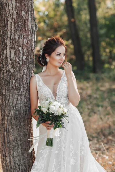 自然の中で結婚式の写真 花嫁は美しいドレスで木の近くに立っている 白いバラの彼女の花束を保持し 側を探している 花嫁の美しいメイク 肖像画 — ストック写真