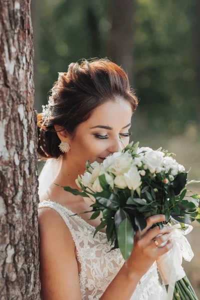 婚纱照在自然界 新娘穿着漂亮的衣服站在一棵树旁 手里拿着一束白玫瑰 望着花束 漂亮的新娘妆容 肖像画 — 图库照片