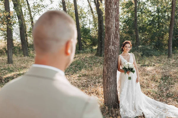 婚纱照在自然界 新娘穿着漂亮的衣服站在一棵树旁 手里拿着一束白玫瑰 望着新郎 在森林里见面 — 图库照片