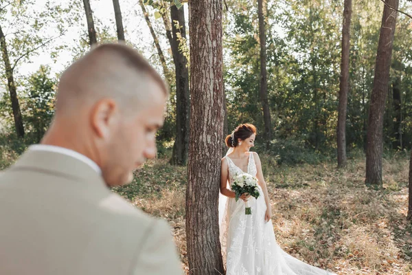 婚纱照在自然界 新娘穿着漂亮的衣服站在一棵树旁 手里拿着一束白玫瑰朝下看 新郎在前面的位置上 — 图库照片