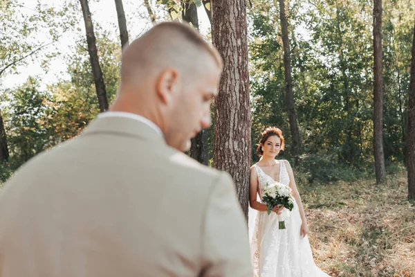 自然の中で結婚式の写真 花嫁は美しいドレスで木の近くに立っている 白いバラの花束を持ち カメラを見ている 新郎は前景にあります — ストック写真