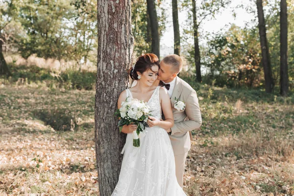 婚纱照在自然界 新娘和新郎站在一棵树旁 新郎从背后拥抱着他心爱的人亲吻她 她真诚地微笑着 夏天的婚礼 — 图库照片