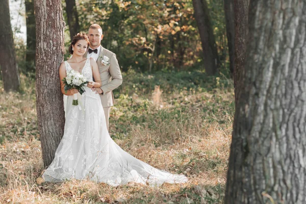 婚纱照在自然界 新娘和新郎笑着站在一棵树旁 看着摄像机 新郎从后面拥抱着他心爱的人 新娘捧着一束花 夏天的婚礼 — 图库照片