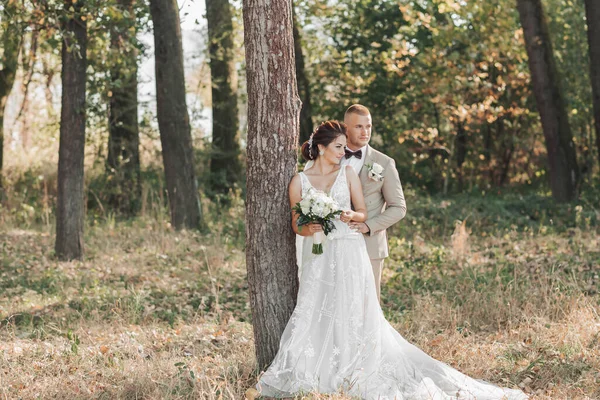 婚纱照在自然界 新娘和新郎笑着站在一棵树旁 望着远方 新郎从后面拥抱着他心爱的人 新娘捧着一束花 夏天的婚礼 — 图库照片