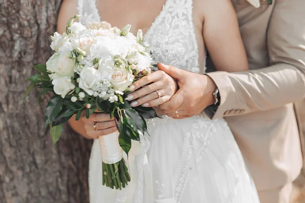 新娘和新郎手中拿着一束白花和绿叶的婚礼花的照片 新郎握住新娘的手 漂亮的手法国修指甲 结婚戒指 — 图库照片