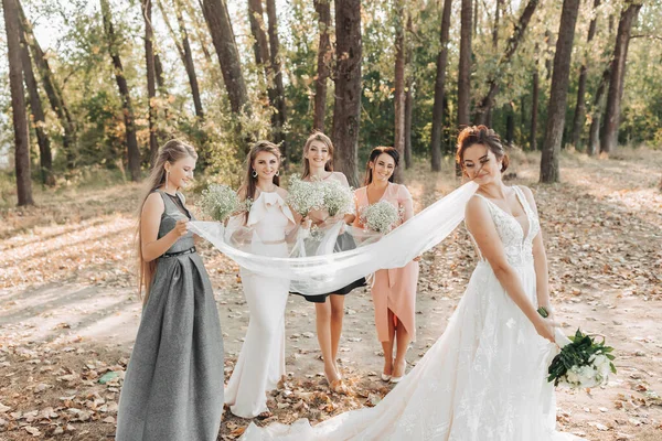 自然の中で結婚式の写真 新婦付添人は花嫁の後ろに立ち 花束および彼女のベールを保持し 花嫁は花束を保持し 見下ろす 結婚式の肖像画 感情だ 美しい女の子 — ストック写真