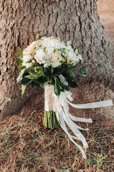 一束由白玫瑰和绿叶组成的婚礼花束 装饰着丝带 矗立在一棵树的背景上 婚礼的细节 新娘的花束 — 图库照片