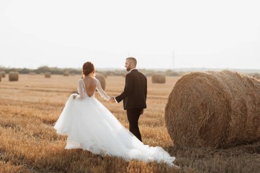 Gelin ve damadın arka planda yürüyen düğün portresi. Damat gelinin elini tutar ve ona bakar, bakışları yere bakar. Uzun elbiseli kızıl saçlı gelin. Şık damat. Yaz