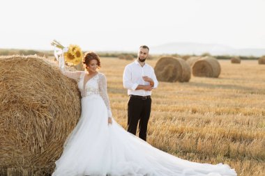 Gelin ve damadın düğün portresi. Damat, gömleğini yırtarak gelinin arkasında duruyor, saman balyasının yanında. Üzerinde bir buket ayçiçeği olan uzun elbiseli kızıl saçlı gelin. Şık damat. Yaz