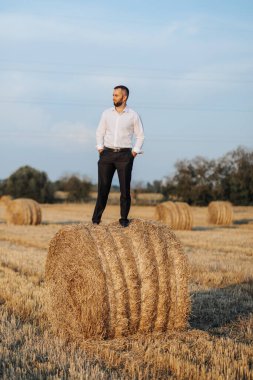 Düğün portresi fotoğrafı. Beyaz gömlekli şık bir damat, elleri cebinde saman balyasında poz veriyor. Sakallı adam. Biçim.