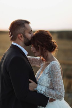 Gelin ve damadın düğün portresi. Damat gelini nazikçe alnından öper. Uzun elbiseli kızıl saçlı gelin. Şık, sakallı damat. Yaz