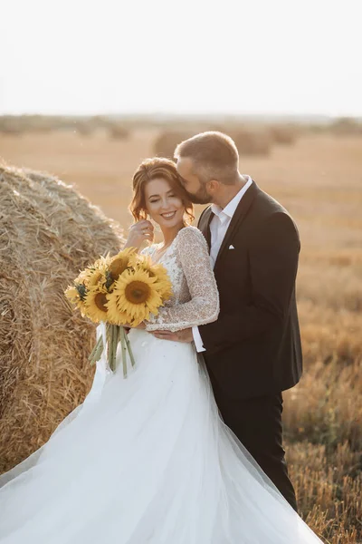 Wedding portrait of the bride and groom. The groom hugs the bride from behind, next to a bale of hay. Side view. Red-haired bride in a long dress with a bouquet of sunflowers. Stylish groom. Summer