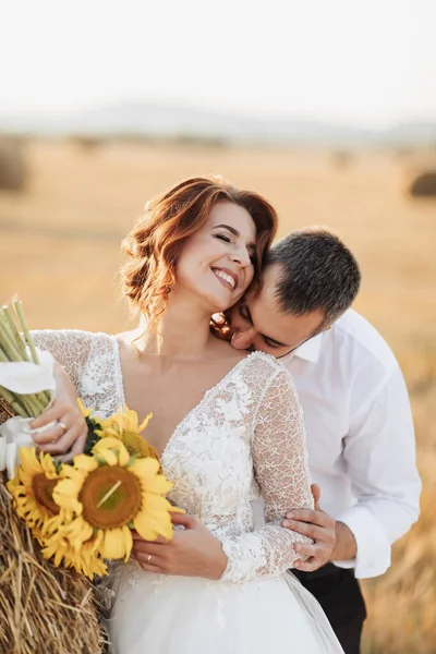 Wedding portrait of the bride and groom. The groom, tearing his shirt, stands behind the bride, near a bale of hay. Red-haired bride in a long dress with a bouquet of sunflowers. Stylish groom. Summer