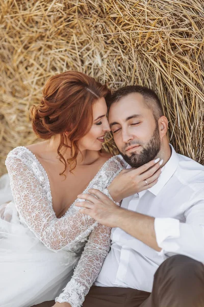 結婚式の肖像画 花嫁と新郎は彼らの目を地面に抱擁では 干し草のベールの近くに閉じて座っている 赤い髪の花嫁の長いドレスです スタイリッシュな新郎 — ストック写真
