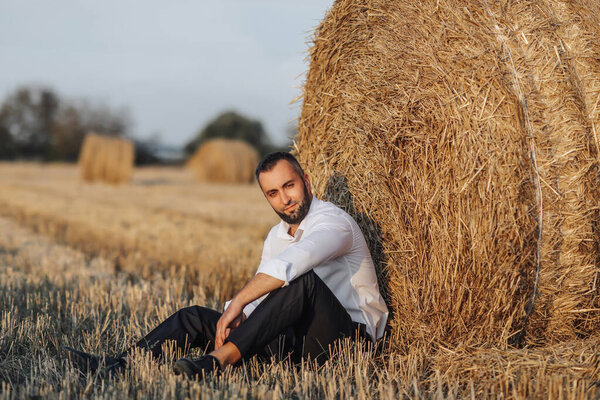 Wedding portrait photo. A stylish groom in a white shirt sits relaxed near a hay bale and looks into the lens. Bearded man. Style.