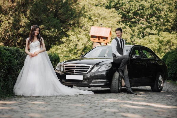 gorgeous bride with trendy makeup and hairdo in luxury wedding dress with handsome groom near black wedding car